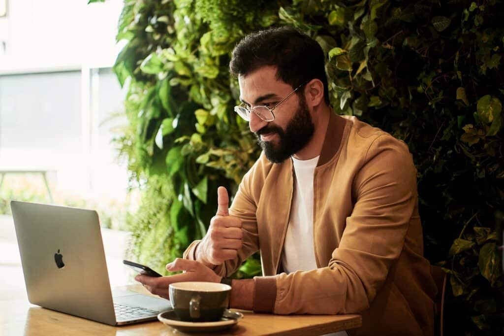 Man in a brown jacket sits at a table and gives a "thumbs-up" to someone on his laptop screen